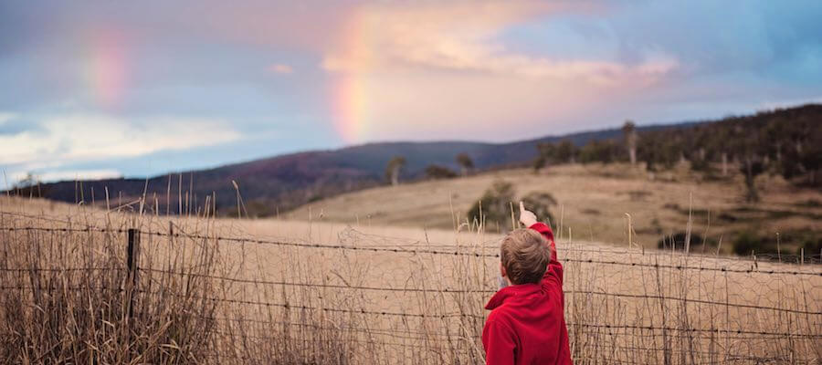 Boy blocked by fence pointing at distant rainbow
